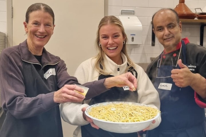Three people holding a large dish of noodles in a kitchen.