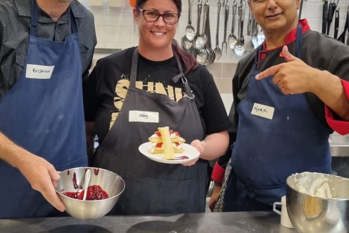 Three people in aprons cooking with a plate of food and tray of churros in a kitchen.