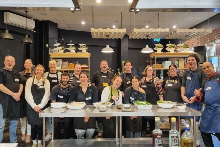 Group of people in aprons posing in a kitchen with prepared dishes on a counter.