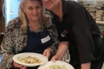Two people smiling and holding plates of pasta in a kitchen setting.