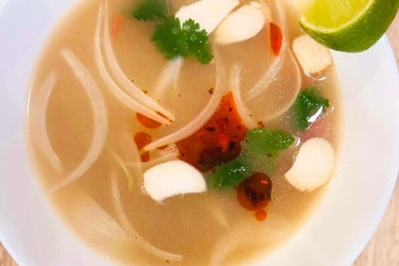 A bowl of soup with lime, cilantro, onions, and white pieces, drizzled with chili oil.