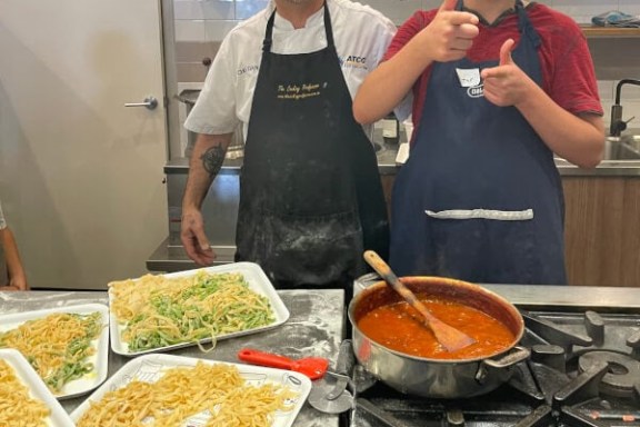 Two people in aprons, one giving thumbs up, stand by trays of pasta and a pot of sauce in a kitchen.