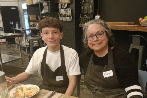 Two people wearing aprons sit at a table with plates of food in a cozy kitchen.