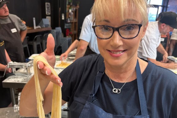 Woman in apron holding fresh pasta with people cooking in the background.