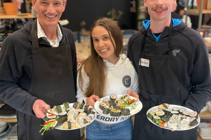 Three people smiling, holding plates of sushi in a kitchen setting.