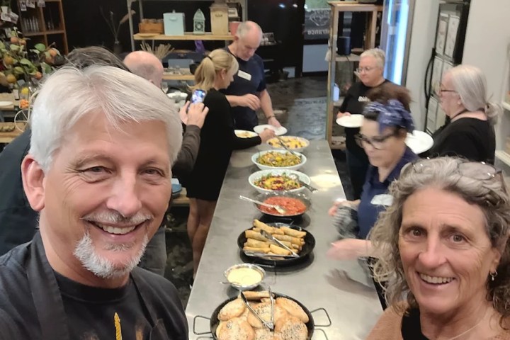 People serving food from a counter; two smiling in foreground, others preparing plates.