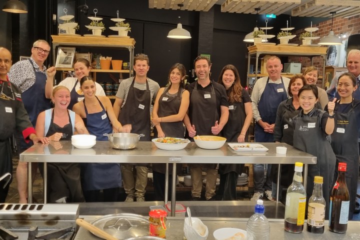 Group of people in aprons posing in a kitchen setting with cooking ingredients on the counter.
