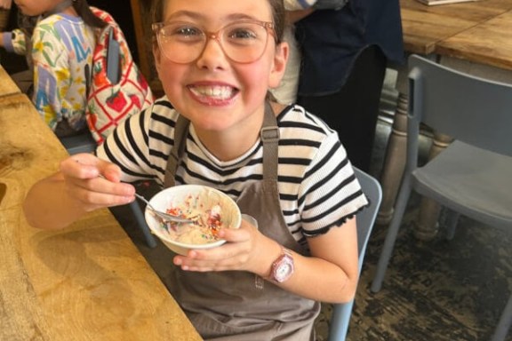 Smiling child in glasses holding a bowl with sprinkles, wearing a striped shirt and apron at a wooden table.