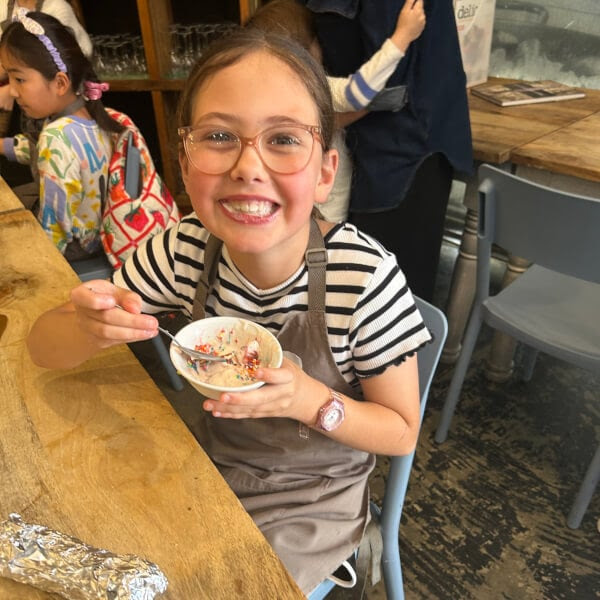 Smiling child in glasses holding a bowl with sprinkles, wearing a striped shirt and apron at a wooden table.