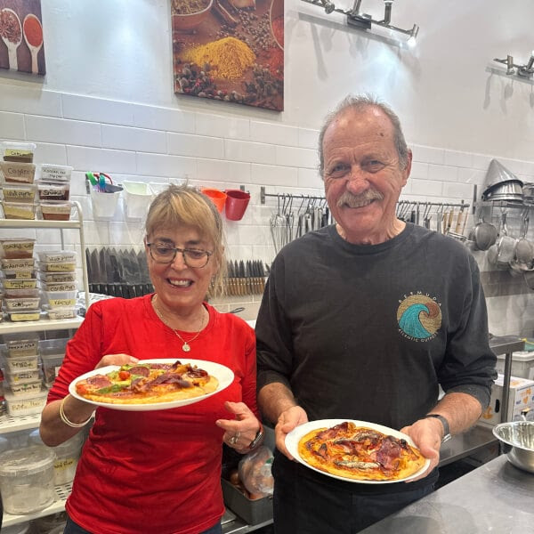 Two people in a kitchen holding plates of pizza, smiling at the camera.