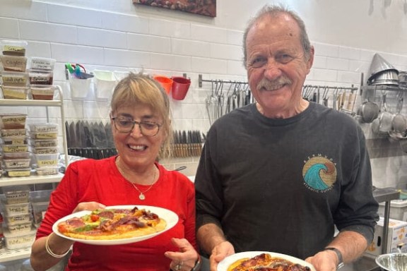 Two people in a kitchen holding plates of pizza, smiling at the camera.
