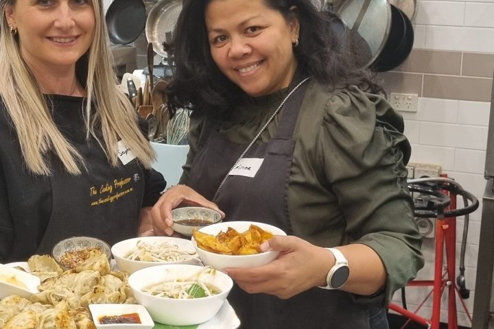 Two women in aprons holding a tray of food in a kitchen setting.