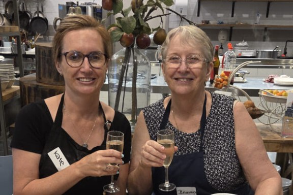 Two women at a kitchen table, holding drinks, surrounded by bowls of food.