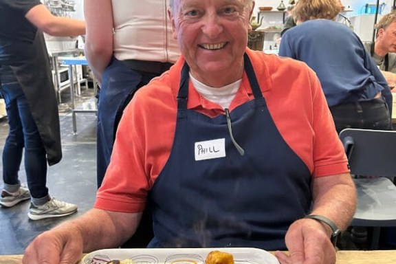 Man in a red shirt and apron holding a plate of food and a bowl of soup in a busy kitchen.