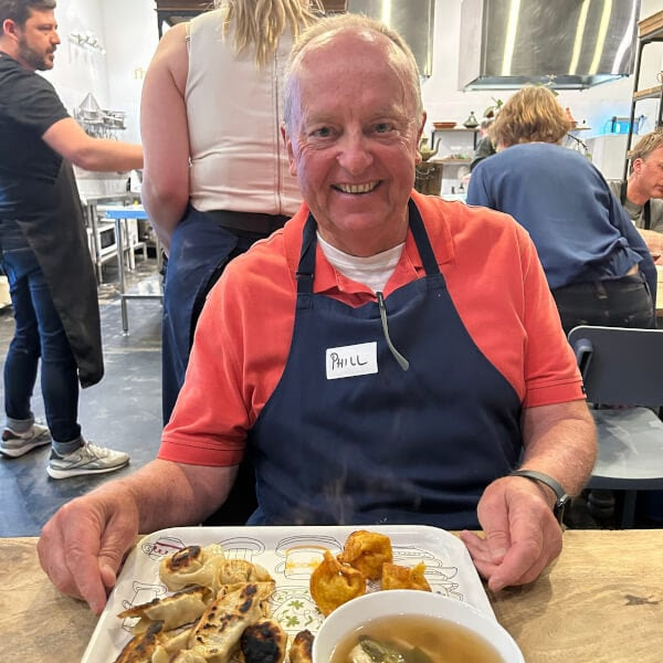 Man in a red shirt and apron holding a plate of food and a bowl of soup in a busy kitchen.