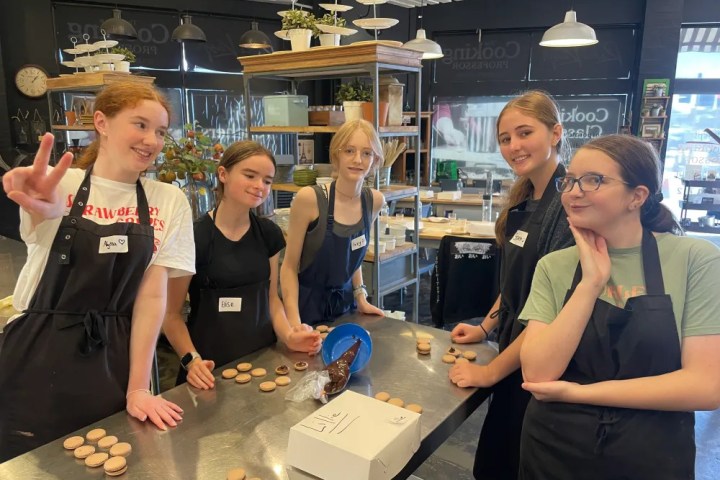 Five people wearing aprons smile while decorating macarons at a kitchen counter.