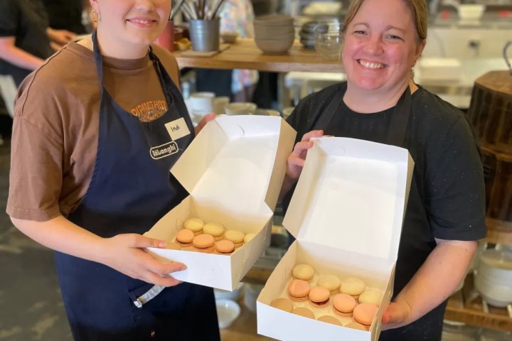 Two women in aprons smiling, holding boxes of macarons in a kitchen setting.