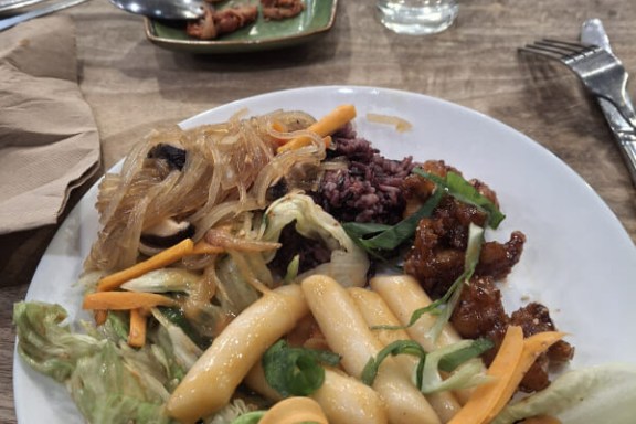 Plate of mixed Korean dishes on a wooden table with utensils and a glass nearby.
