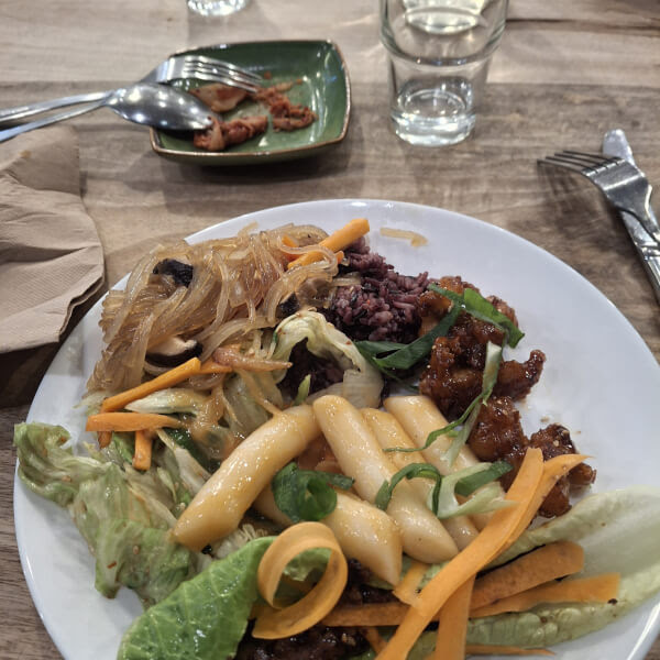 Plate of mixed Korean dishes on a wooden table with utensils and a glass nearby.