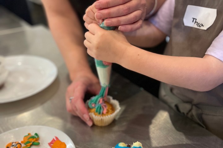 Child and adult decorating cupcakes with multicolored frosting and decorations on table.