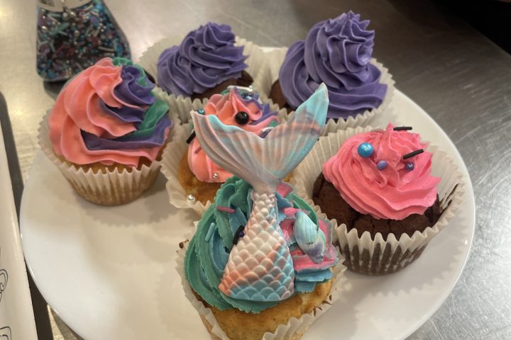Plate of colorful cupcakes with mermaid and sprinkle decorations on a kitchen counter.