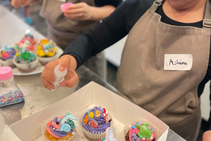 Two children in aprons decorate colorful cupcakes with icing and sprinkles in a kitchen.