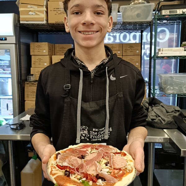 Person smiling, holding a pizza with various toppings in a kitchen setting.