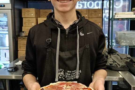 Person smiling, holding a pizza with various toppings in a kitchen setting.