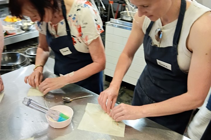 Two women in aprons folding pastry at a kitchen counter.