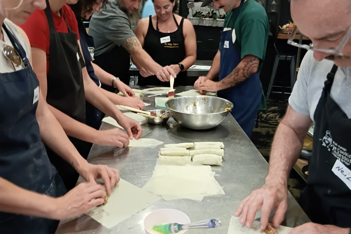 Group of people in aprons preparing food around a stainless steel table.
