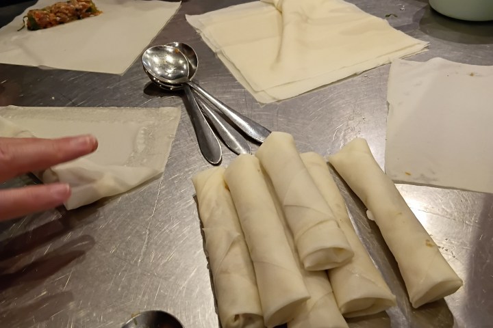Hands rolling spring rolls on a metal table with spoons.