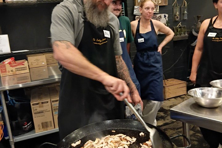 Man cooking sliced meat in a large pan with onlookers wearing aprons.
