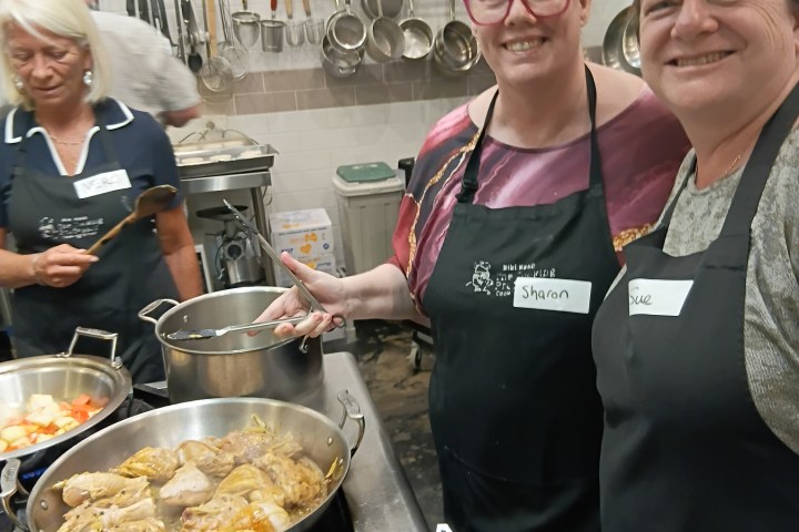 Three people cooking, one stirring a pot, others smiling, in a kitchen with pots and utensils on the wall.