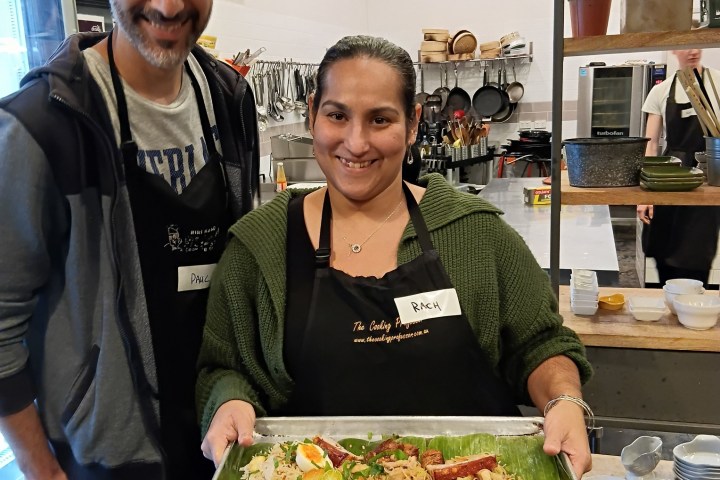 Two people smiling, one holding a tray with various dishes in a kitchen setting.