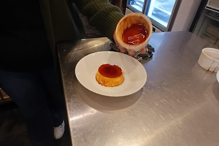 Person holding dessert plate with caramel flan in a kitchen.