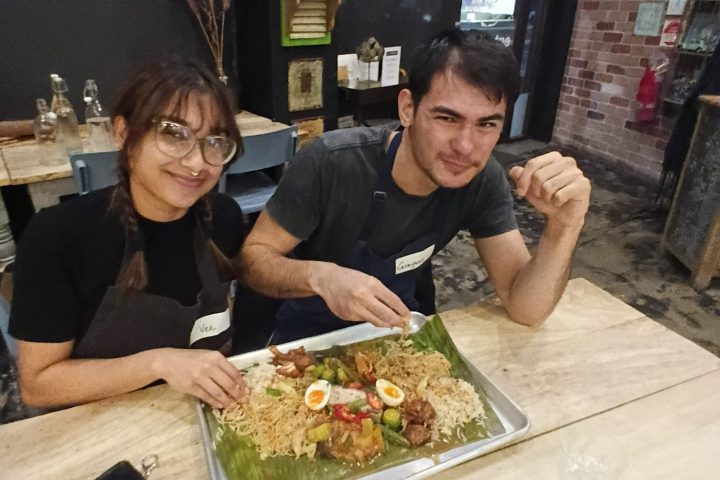 Two people smiling at a table with a large dish of food on banana leaves.