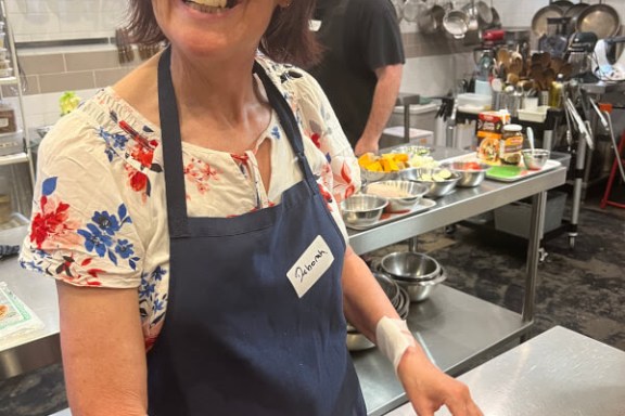 Smiling woman in a kitchen wearing blue apron, preparing food on a metal counter.