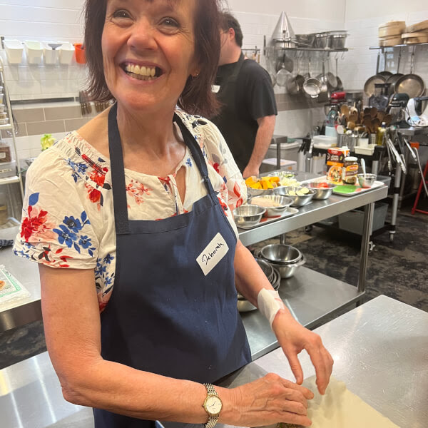 Smiling woman in a kitchen wearing blue apron, preparing food on a metal counter.