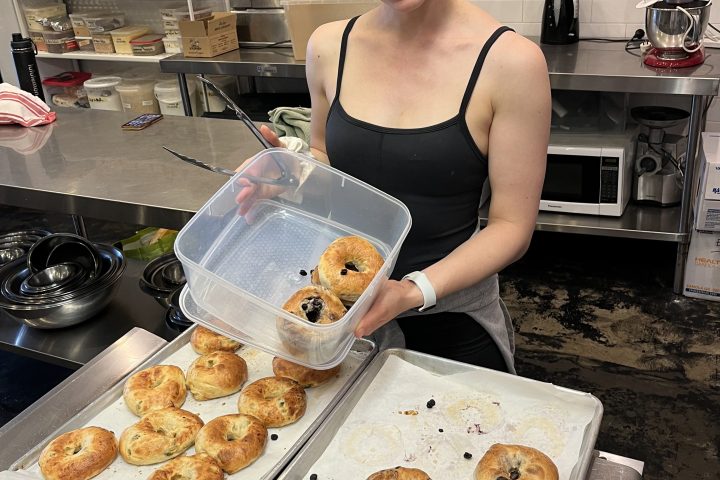 Smiling woman holding tray of bagels in a kitchen.