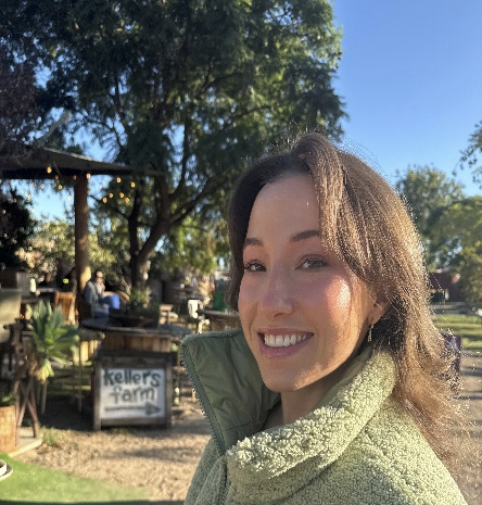 Woman smiling outdoors at a farm with trees and a sign saying 'Kellers Farm'