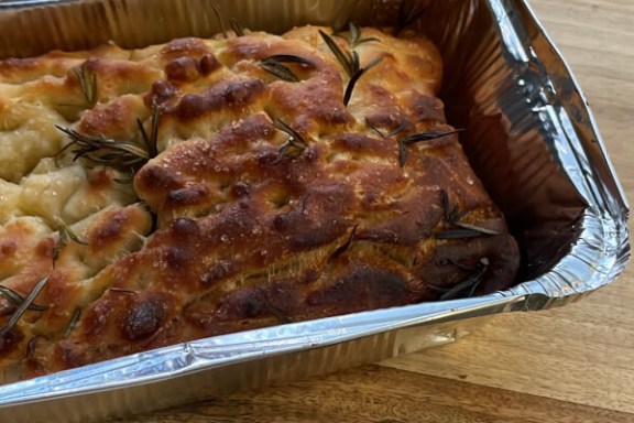 Baked focaccia with rosemary sprigs in a foil tray on a wooden surface.