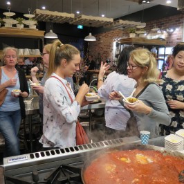 A group of people tasting food near a large pot of simmering tomato sauce in a cooking class setting.