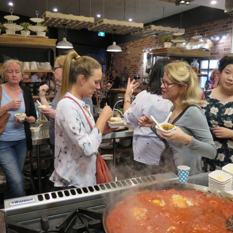 A group of people tasting food near a large pot of simmering tomato sauce in a cooking class setting.