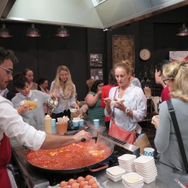 People gathered around a large pan of food, some serving themselves and others eating.