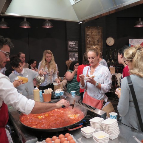People gathered around a large pan of food, some serving themselves and others eating.