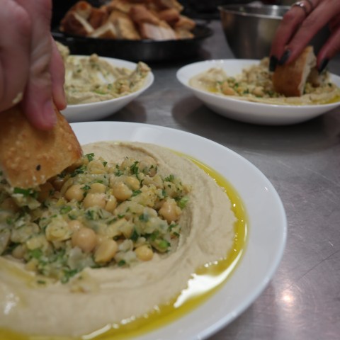 Hands dipping bread into hummus topped with chickpeas and herbs on a plate.