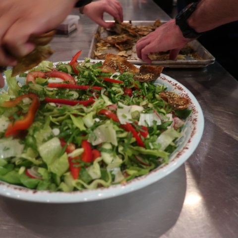 Hands preparing salad in a bowl with mixed greens and sliced red peppers on a counter.