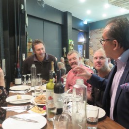 Group of people dining, one man standing and gesturing while others listen.