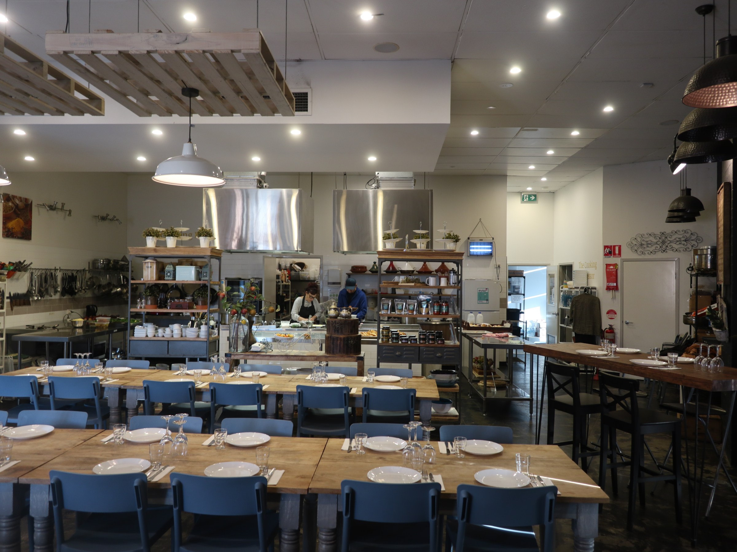 Spacious kitchen with long tables, blue chairs, and chefs preparing food in the background.