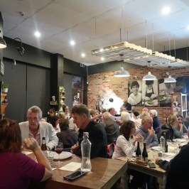 A crowded restaurant with people sitting at tables, talking and eating, with a brick wall and photos in the background.